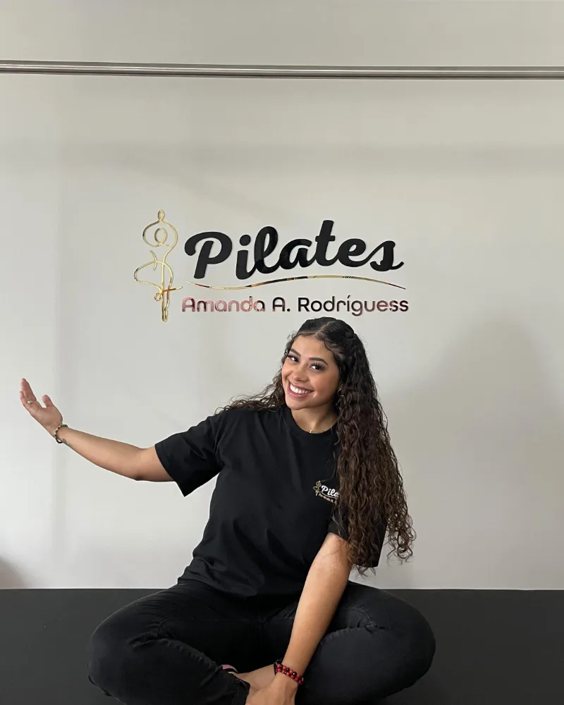 Amanda sentada no studio, em frente à parede com o logo Pilates Amanda A. Rodríguess